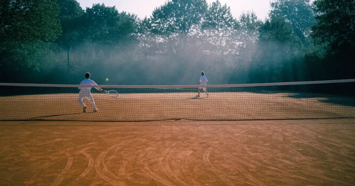 Ein Tennisplatz aus Sand bei Sonnenaufgang mit gespanntem Netz, Nebel zwischen den Bäumen am Rand