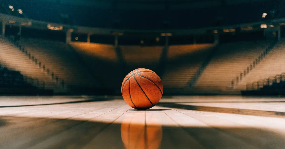 Ein Basketball liegt auf dem hellen Parkett einer leeren Halle, im Hintergrund steigen die Tribünenreihen im Halbdunkel an.