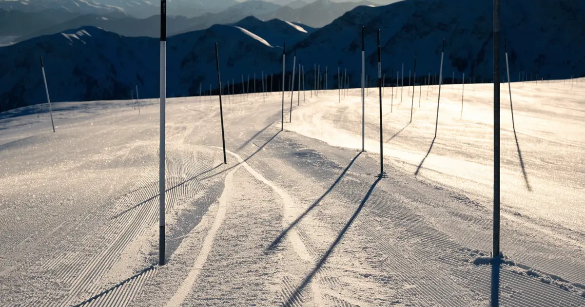 Ein frisch präparierter Hang mit Slalomstangen in einer Reihe im ersten Morgenlicht, Schneespuren im Hang und Berge im Hintergrund.