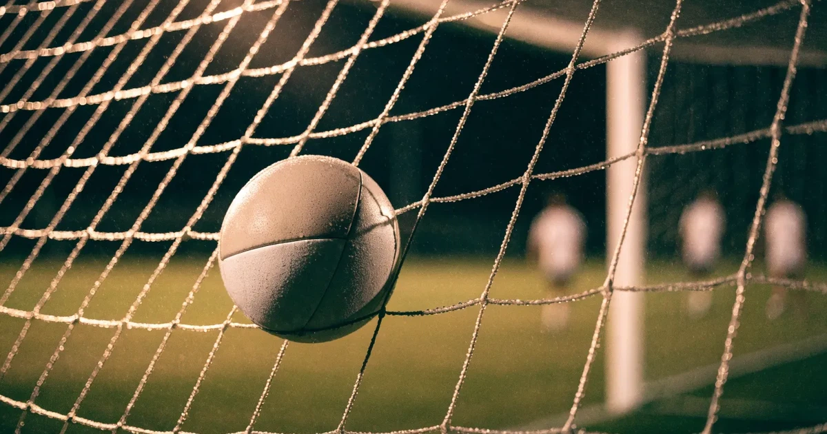 Ein Fussballnetz in Nahaufnahme, in dem ein Ball hängt, Wassertropfen glänzen auf den Maschen.