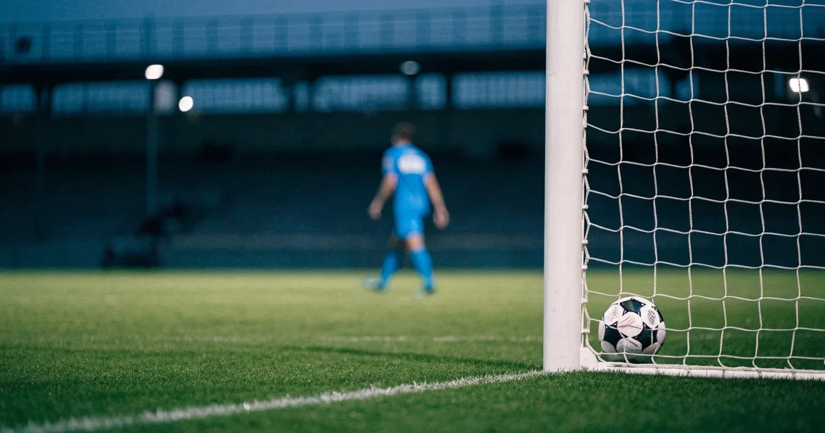 Ein Fussball liegt im Netz eines Tores, Flutlicht und kalte Abendluft liegen über dem leeren Platz